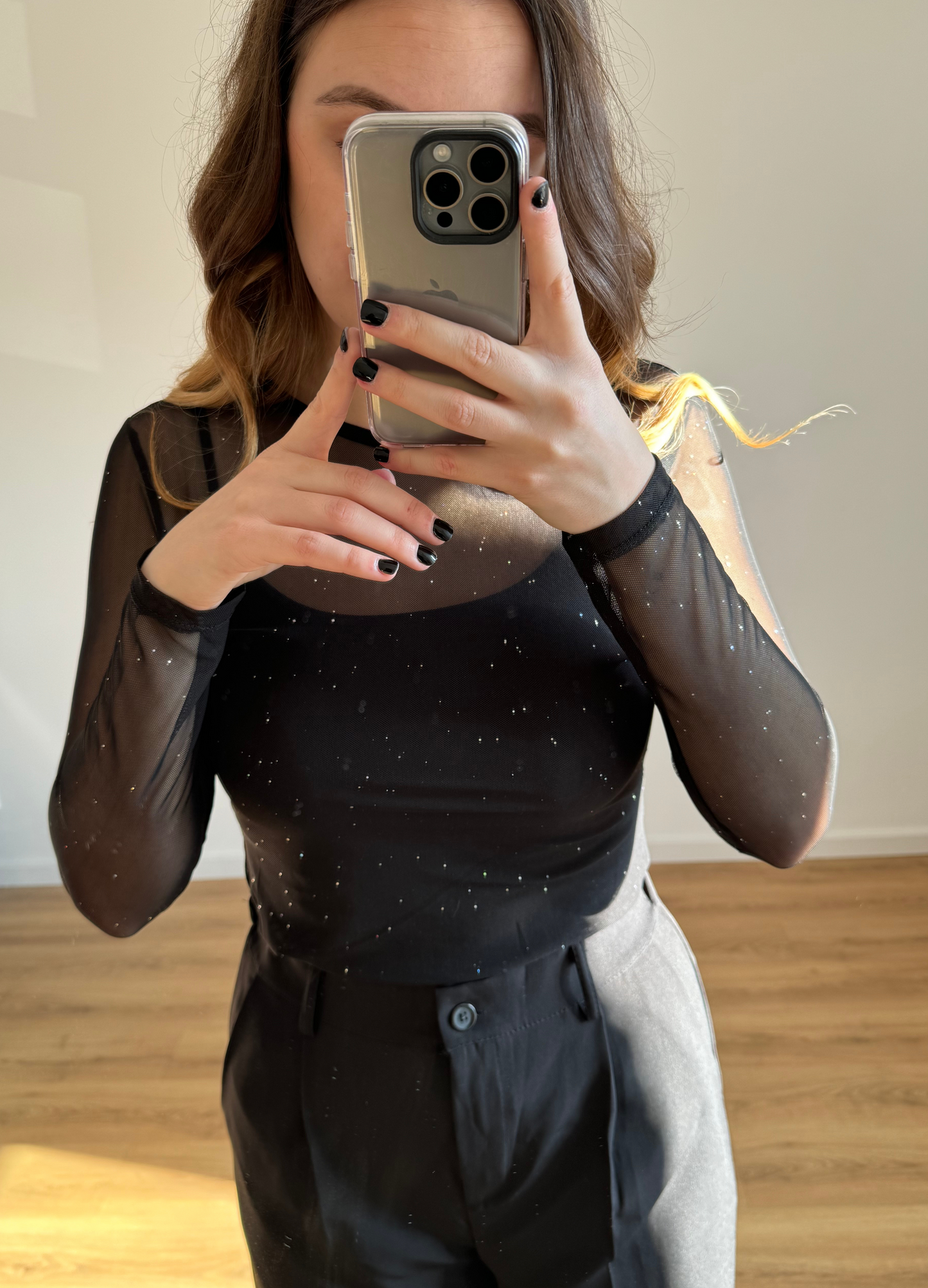 Femme portant un élégant haut noir en tulle à manches longues orné de détails brillants, assorti à un pantalon noir taille haute et ample. Look chic et moderne, idéal pour une tenue de soirée ou un événement habillé.