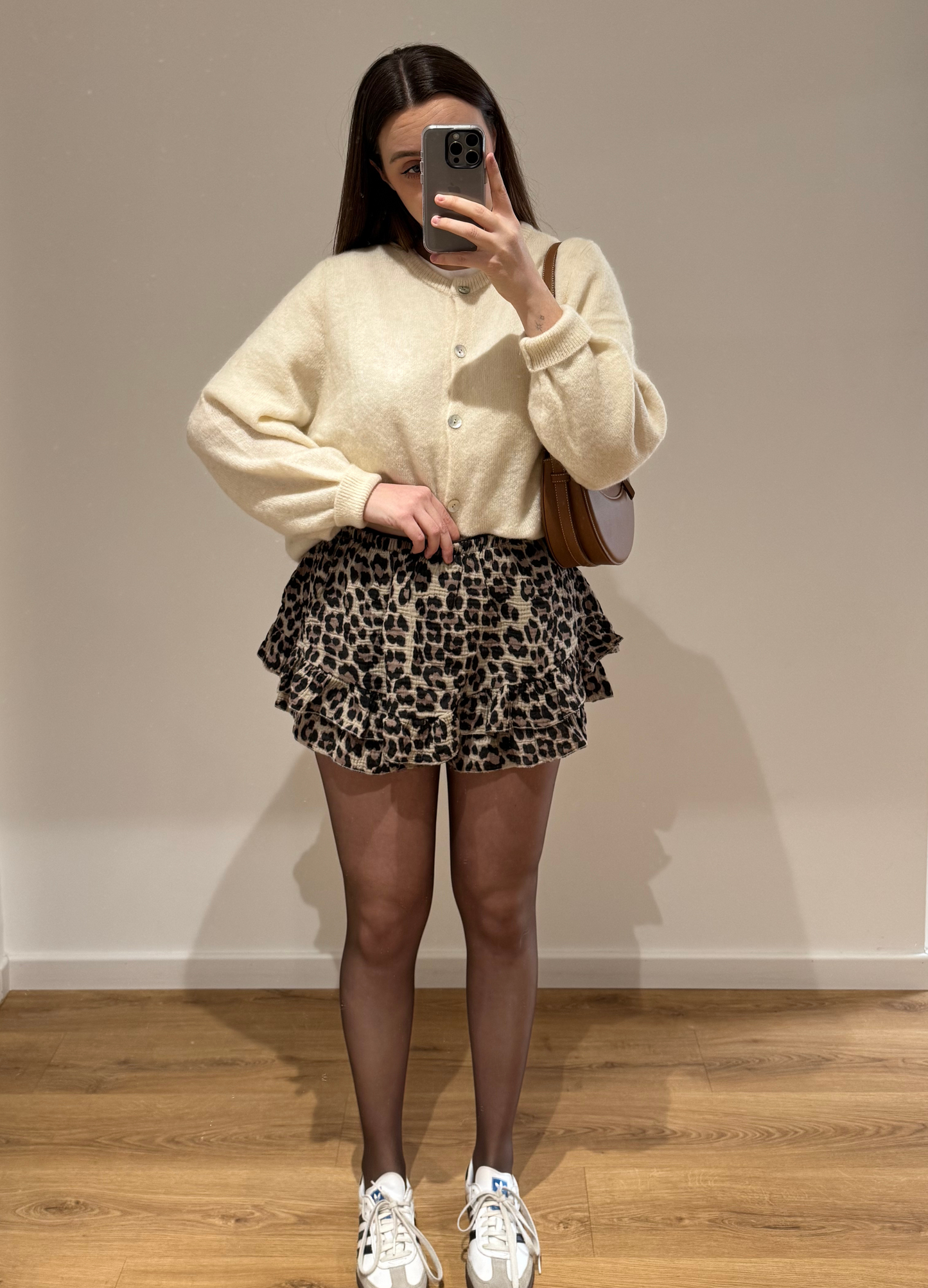 Femme portant un cardigan blanc OLIVIA en coton à boutons argenté, avec un short à motifs léopard KLEOFINA , vue en intérieur sur parquet et fond blanc.