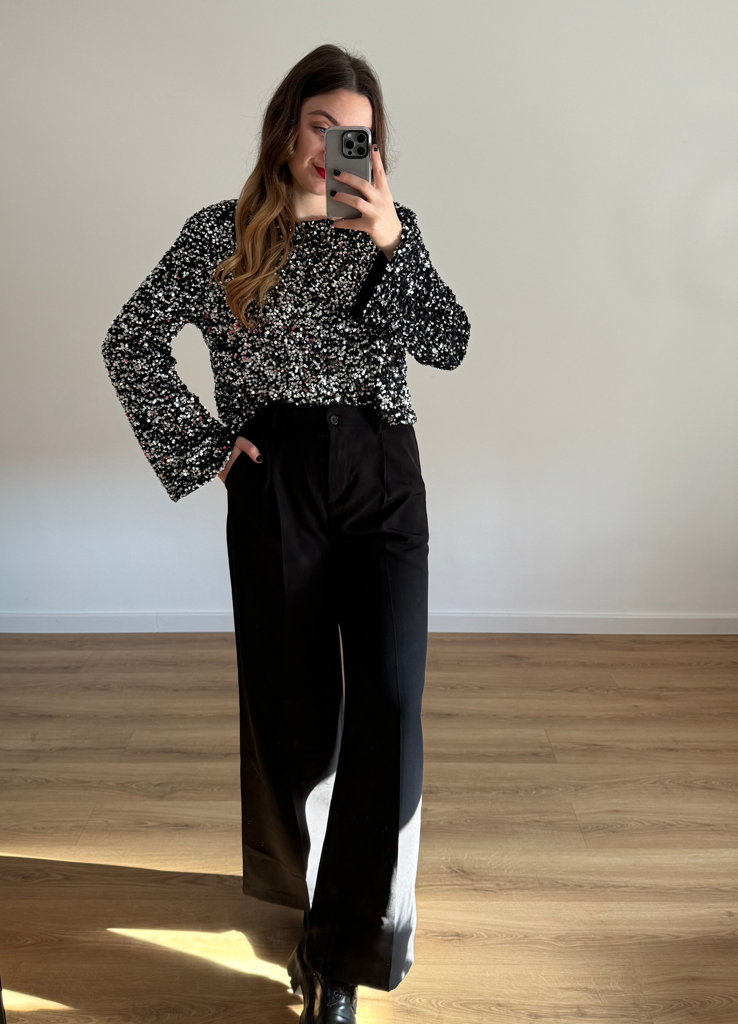 Femme prenant un selfie tout en portant un haut en velour noir à stras à manches longues, associé à un pantalon noir taille haute et ample. Look élégant et intemporel, idéal pour une tenue habillée ou casual chic.