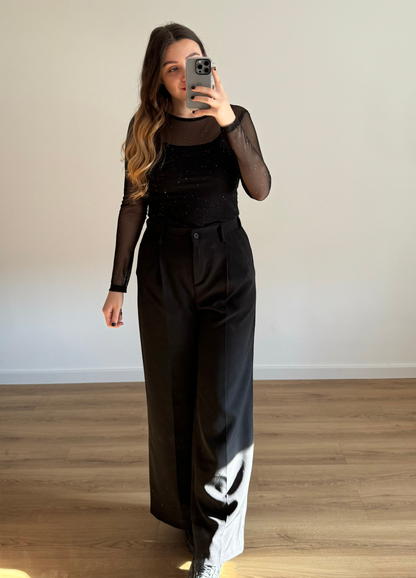 Femme portant un élégant haut noir en tulle à manches longues orné de détails brillants, assorti à un pantalon noir taille haute et ample. Look chic et moderne, idéal pour une tenue de soirée ou un événement habillé. 