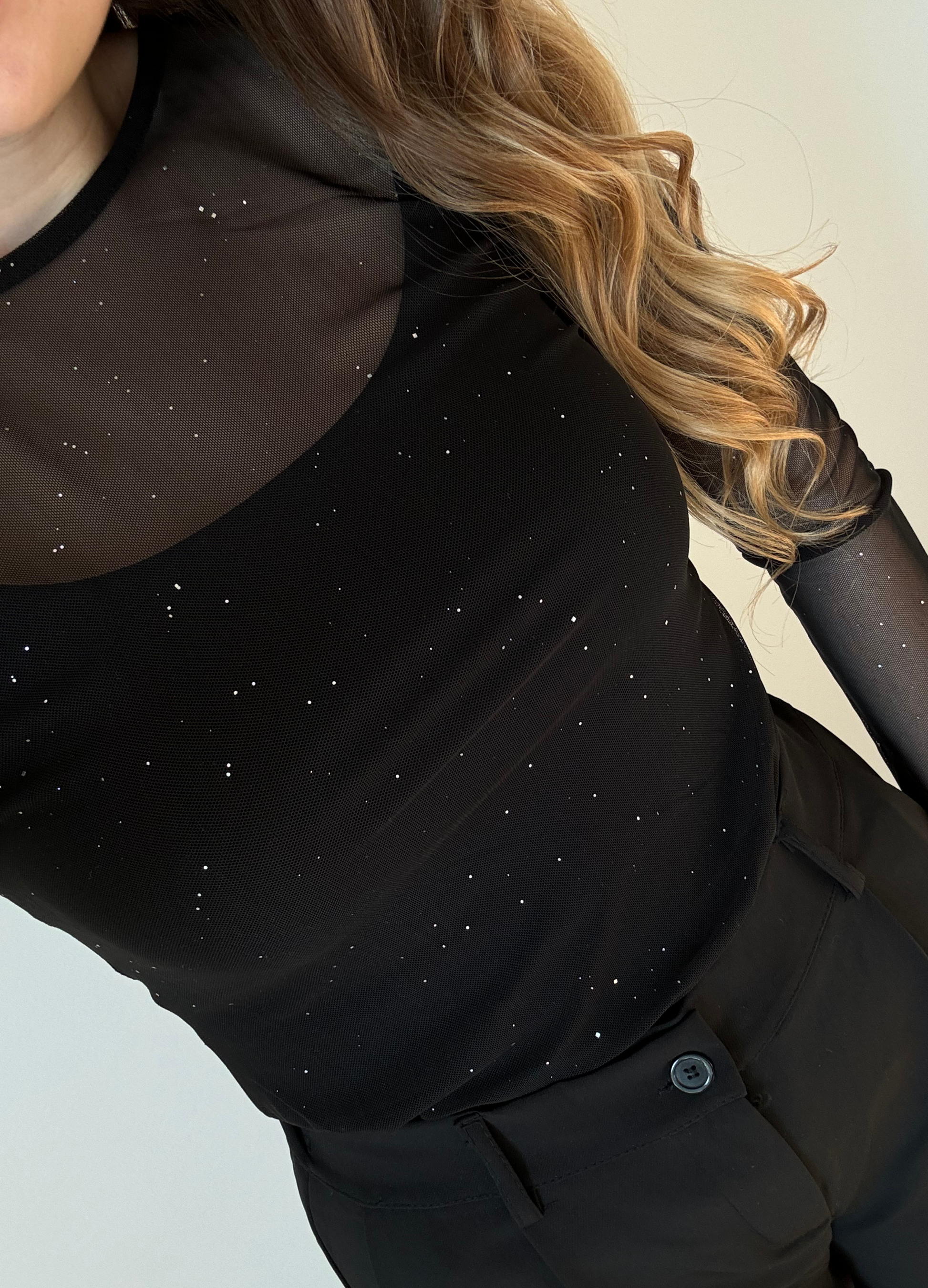 Femme portant un élégant haut noir transparent à manches longues orné de détails brillants, assorti à un pantalon noir taille haute et ample. Look chic et moderne, idéal pour une tenue de soirée ou un événement habillé.